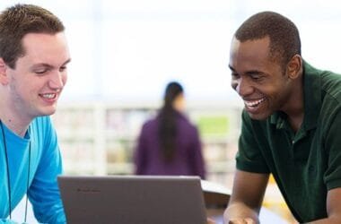 Homem de pele preta e camisa verde musgo interage com vendedor da Microsoft, um homem branco usando camisa azul claro, enquanto olham para um notebook na loja da marca