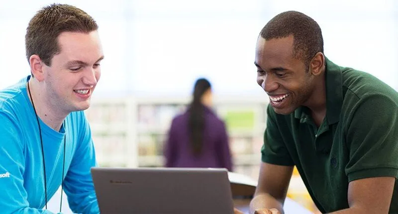 Homem de pele preta e camisa verde musgo interage com vendedor da Microsoft, um homem branco usando camisa azul claro, enquanto olham para um notebook na loja da marca