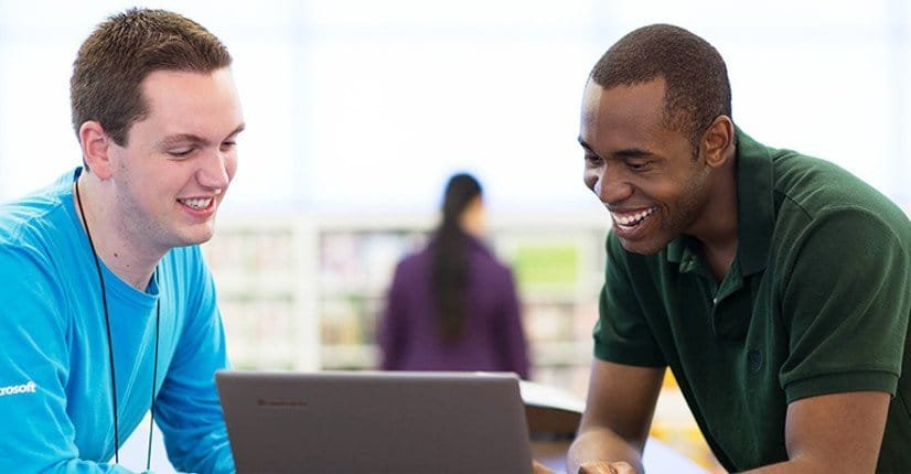 Homem de pele preta e camisa verde musgo interage com vendedor da Microsoft, um homem branco usando camisa azul claro, enquanto olham para um notebook na loja da marca