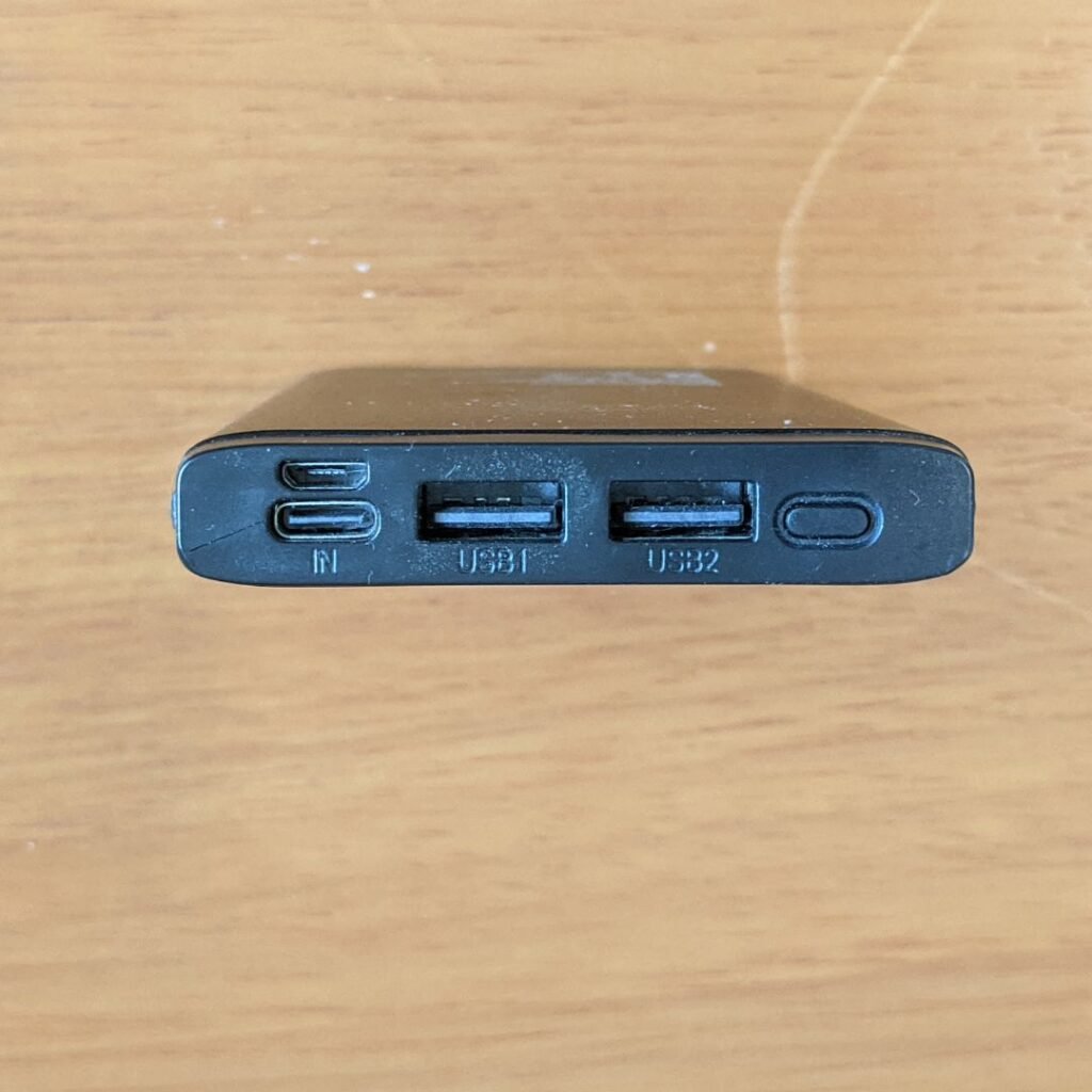 A imagem mostra a parte superior de um power bank Lity Chrono CRK-100 deitado sobre uma superfície de madeira clara. As portas do power bank estão visíveis e rotuladas da seguinte forma: uma porta USB-C e uma micro USB rotuladas como "IN", uma porta USB-A rotulada como "USB1", uma porta USB-A rotulada como "USB2" e uma botão oval para ligar e desligar a bateria portátil.