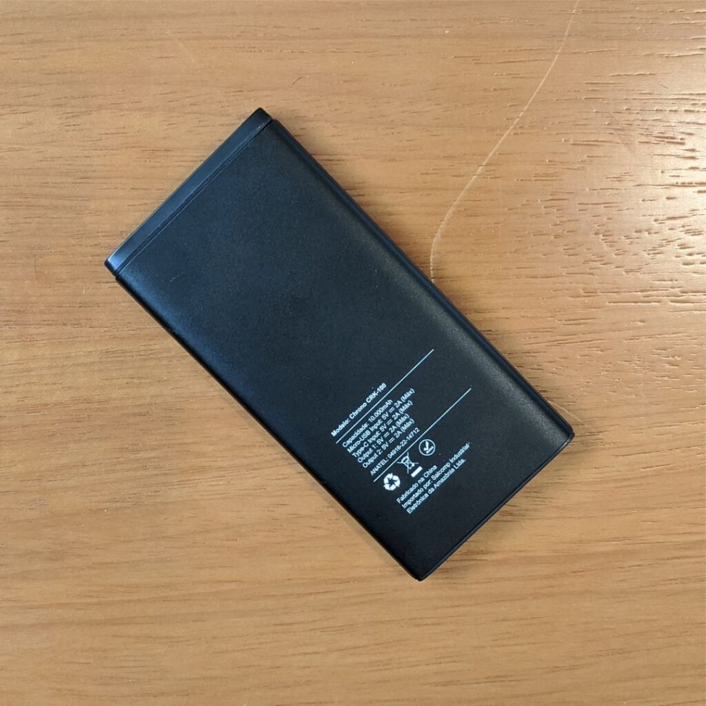 Esta imagem apresenta um power bank Lity Chrono CRK-100 visto de cima, em um ângulo ligeiramente inclinado. O power bank é preto e retangular, com informações impressas na parte de trás. A etiqueta na parte de trás inclui o modelo, capacidade, especificações de entrada e saída, e informações do fabricante. O power bank está sobre uma mesa de madeira.