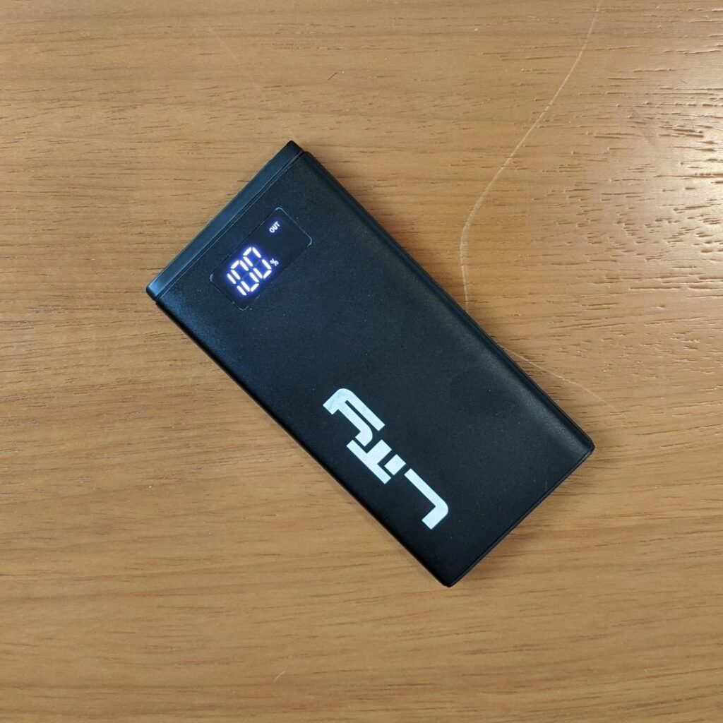 A imagem mostra um power bank Lity Chrono CRK-100 visto de cima, em um ângulo ligeiramente inclinado. O power bank é preto e retangular, com o logotipo "Lity" impresso na parte inferior. Há um display digital na parte superior que mostra "100%" e a palavra "OUT". O power bank está sobre uma mesa de madeira clara.