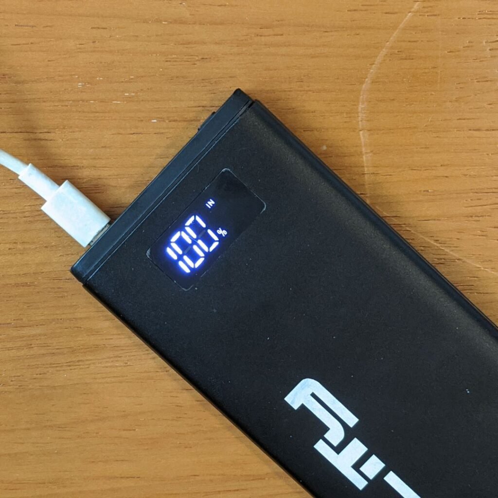 A imagem mostra um power bank Lity Chrono CRK-100 visto de cima, em um ângulo próximo. O power bank é preto e retangular, com o logotipo "Lity" impresso na parte inferior. Há um display digital na parte superior que mostra "100%" e a palavra "IN". Um cabo branco está conectado à porta de carregamento do power bank, localizada na parte superior. O power bank está sobre uma mesa de madeira clara.