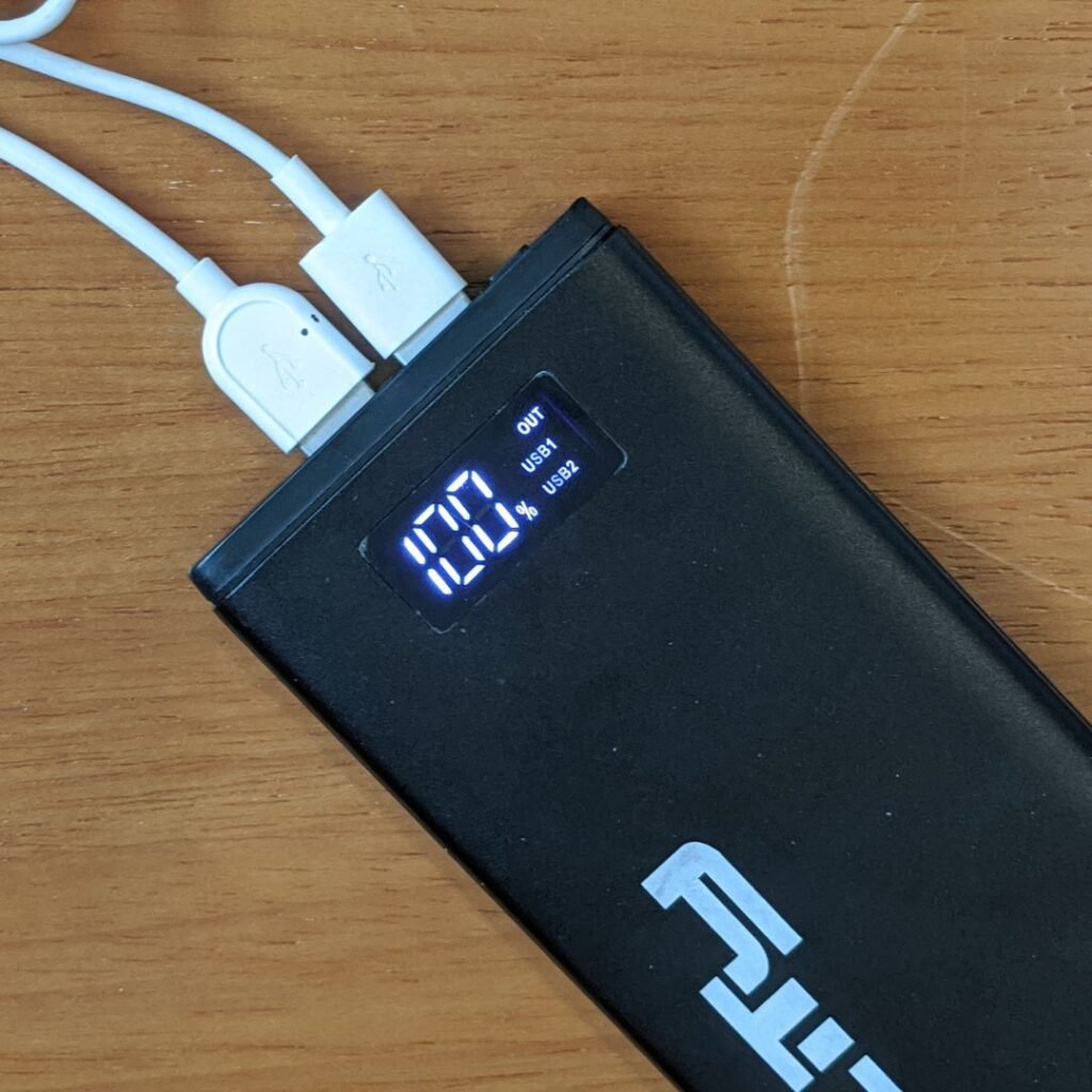 A imagem mostra um power bank Lity Chrono CRK-100 visto de cima, em um ângulo próximo. O power bank é preto e retangular, com o logotipo "Lity" impresso na parte inferior. Há um display digital na parte superior que mostra "100%" e as palavras "OUT", "USB1" e "USB2". Dois cabos brancos estão conectados às portas USB-A do power bank, localizadas na parte superior. O power bank está sobre uma mesa de madeira clara.
