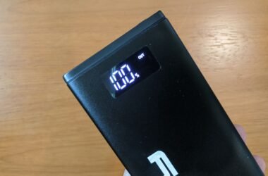 A imagem mostra um power bank Lity Chrono CRK-100 sendo segurado contra um fundo de madeira. O power bank é preto e retangular, com um display digital iluminado que mostra "100%" e a palavra "OUT".