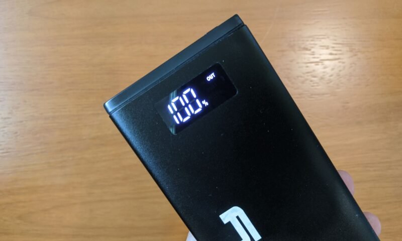 A imagem mostra um power bank Lity Chrono CRK-100 sendo segurado contra um fundo de madeira. O power bank é preto e retangular, com um display digital iluminado que mostra "100%" e a palavra "OUT".