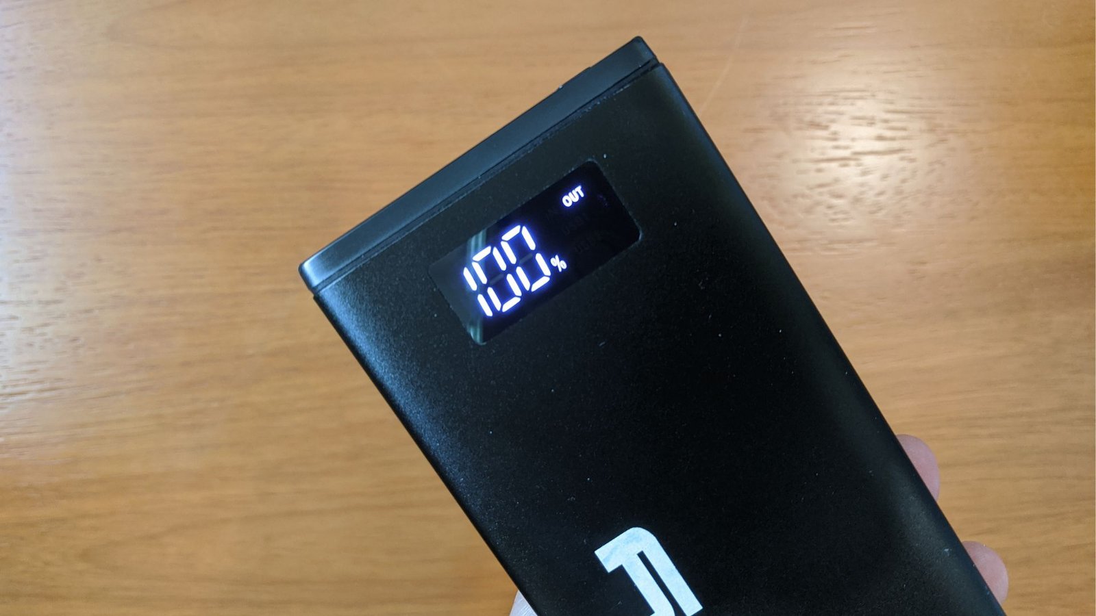 A imagem mostra um power bank Lity Chrono CRK-100 sendo segurado contra um fundo de madeira. O power bank é preto e retangular, com um display digital iluminado que mostra "100%" e a palavra "OUT".