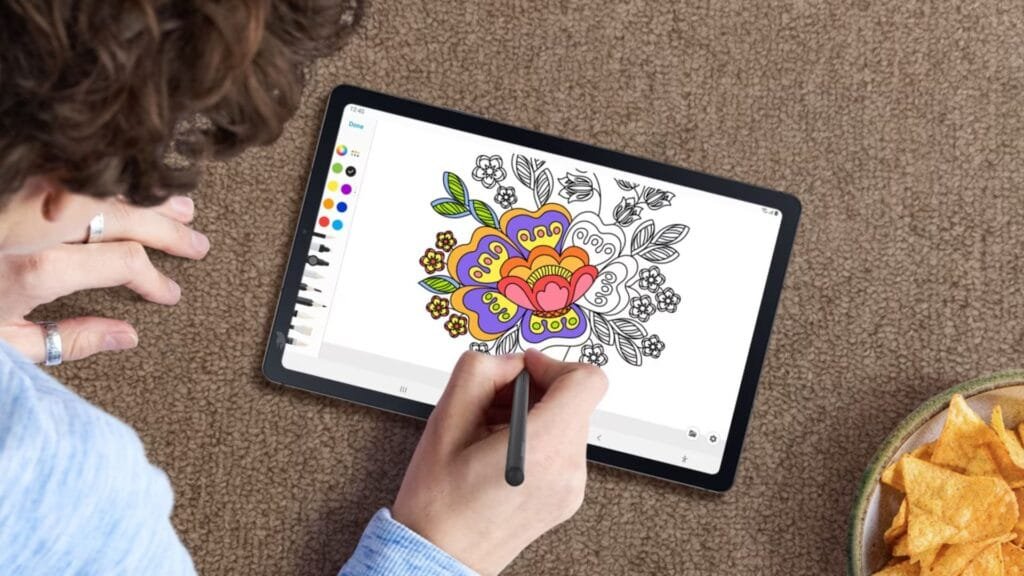Imagem exibe uma pessoa branca com cabelos castanhos e cacheados usando um agasalho mescla entre azul e branco com dois anéis prateados no polegar e dedo do meio usando uma S Pen para colorir uma flor no Samsung Galaxy Tab S6 Lite, que aparece centralizado na imagem. No lado direito, é possível perceber em segundo plano uma tigela com batatas fritas em formato chip