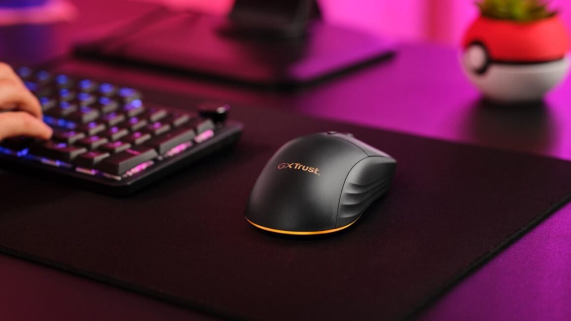 Um mouse gamer sem fio Trust Gaming GXT 980 Redex preto é visto de cima e de lado, sobre um mousepad preto. A mão de uma pessoa está parcialmente visível, digitando em um teclado gamer. O mouse tem um design ergonômico, com linhas curvas e uma base que se ilumina em laranja (a cor é configurável, mas na imagem aparece laranja). O logotipo "GXT Trust" é visto no mouse. O mouse está em um ambiente de jogo, com um teclado mecânico parcialmente visível ao fundo, e iluminação ambiente roxa e rosa. É importante realçar que ele é sem fio e recarregável.