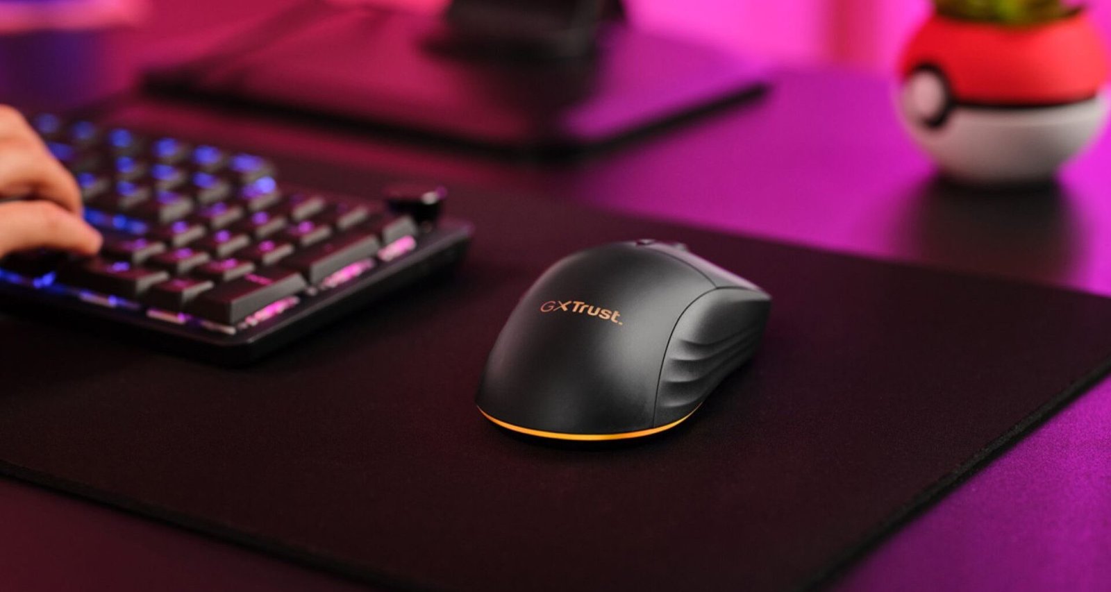 Um mouse gamer sem fio Trust Gaming GXT 980 Redex preto é visto de cima e de lado, sobre um mousepad preto. A mão de uma pessoa está parcialmente visível, digitando em um teclado gamer. O mouse tem um design ergonômico, com linhas curvas e uma base que se ilumina em laranja (a cor é configurável, mas na imagem aparece laranja). O logotipo "GXT Trust" é visto no mouse. O mouse está em um ambiente de jogo, com um teclado mecânico parcialmente visível ao fundo, e iluminação ambiente roxa e rosa. É importante realçar que ele é sem fio e recarregável.