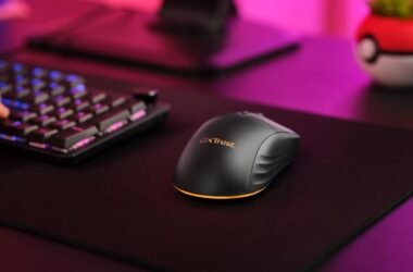 Um mouse gamer sem fio Trust Gaming GXT 980 Redex preto é visto de cima e de lado, sobre um mousepad preto. A mão de uma pessoa está parcialmente visível, digitando em um teclado gamer. O mouse tem um design ergonômico, com linhas curvas e uma base que se ilumina em laranja (a cor é configurável, mas na imagem aparece laranja). O logotipo "GXT Trust" é visto no mouse. O mouse está em um ambiente de jogo, com um teclado mecânico parcialmente visível ao fundo, e iluminação ambiente roxa e rosa. É importante realçar que ele é sem fio e recarregável.