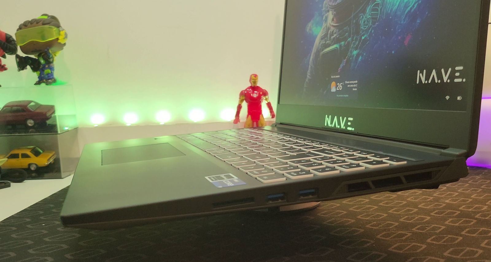 Visão lateral em perspectiva de um notebook NAVE GAMER Supernova GM6IX7X, posicionado sobre um suporte giratório de metal prateado. O notebook está ligeiramente elevado, revelando a lateral direita com suas portas de conexão: duas portas USB-A e uma entrada para cartão de memória SD. O teclado retroiluminado em branco e o touchpad são claramente visíveis, assim como a tela com bordas finas, exibindo um papel de parede colorido com a imagem de um personagem. Uma luz verde neon emana do fundo, iluminando a parede atrás do notebook. No canto superior esquerdo, miniaturas de carros e figuras colecionáveis em uma prateleira embaçada sugerem um ambiente de jogos, incluindo uma figura do Homem de Ferro em destaque. O suporte do notebook e a superfície do mousepad texturizado abaixo são iluminados por uma luz roxa suave.
