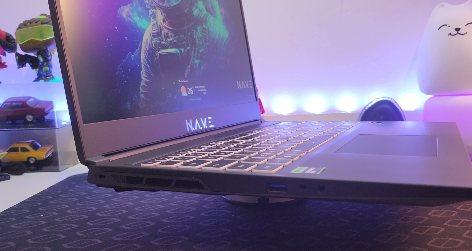 Visão lateral em perspectiva de um notebook NAVE GAMER Supernova GM6IX7X, posicionado sobre um suporte giratório de metal prateado. O notebook está ligeiramente elevado, revelando a lateral esquerda com suas portas de conexão: uma porta USB-A e entrada para fone de ouvido com ponta dupla. O teclado retroiluminado em branco e o touchpad são claramente visíveis, assim como a tela com bordas finas, exibindo um papel de parede colorido com a imagem de um astronauta. Uma luz roxa iluminanda a superfície do mousepad texturizado abaixo. No canto superior direito, um objeto branco com um rosto de gato sorridente é visível, enquanto no canto superior esquerdo, miniaturas de carros e figuras colecionáveis em uma prateleira embaçada sugerem um ambiente de jogos.