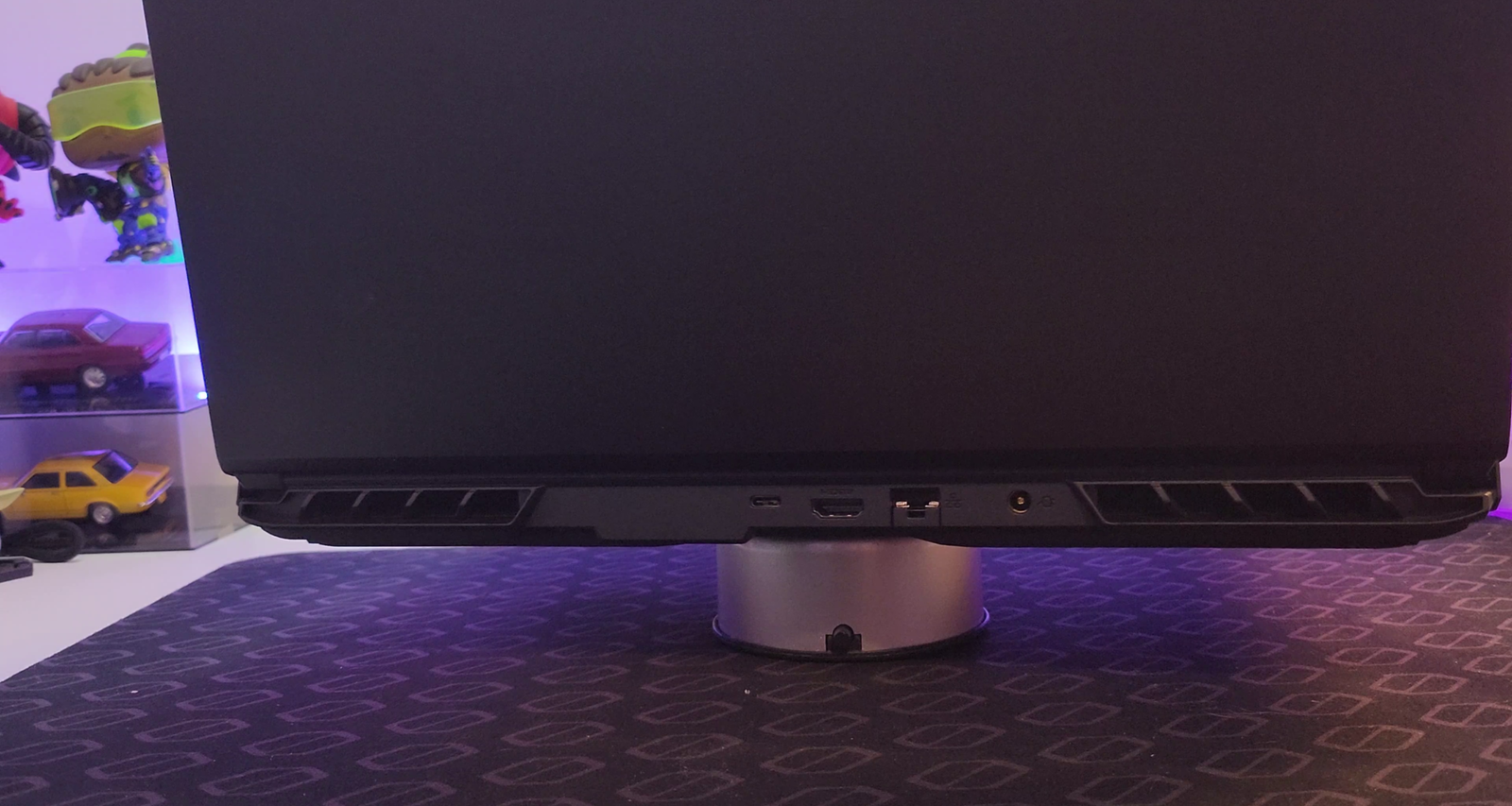 Visão traseira de um notebook preto, o NAVE GAMER Supernova GM6IX7X, posicionado em um suporte circular de metal prateado. O notebook está ligeiramente elevado, mostrando a faixa traseira onde se encontram as portas de conexão: HDMI, USB-C e uma entrada para carregador. As saídas de ar, com design recortado, são visíveis nas extremidades da faixa traseira. A parte inferior do notebook e o suporte são iluminados por uma luz roxa suave. Ao fundo, fora de foco, um mousepad preto texturizado e uma prateleira com objetos coloridos, incluindo miniaturas de carros e figuras colecionáveis, sugerem um ambiente de jogos.