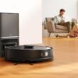 Um robô aspirador de pó preto (TP-Link Tapo RV30 Max Plus) é mostrado em primeiro plano, posicionado na frente de uma estação de acoplamento preta vertical. A estação tem uma pequena luz branca na frente. O robô aspirador tem um disco cinza no centro e um pequeno disco cinza na lateral. Está sobre um piso de madeira bege claro. No fundo, uma família de três pessoas (um homem sentado em um sofá bege com almofadas cinzas e uma menina com uma garota um pouco mais velha estão deitadas no chão. Todos estão usando roupas casuais. O homem parece trabalhar em um notebook enquanto as meninas parecem se divertir desenhando ou escrevendo. O cômodo é bem iluminado com luz natural e apresenta uma parede branca com batente de porta bege.