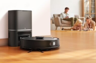 Um robô aspirador de pó preto (TP-Link Tapo RV30 Max Plus) é mostrado em primeiro plano, posicionado na frente de uma estação de acoplamento preta vertical. A estação tem uma pequena luz branca na frente. O robô aspirador tem um disco cinza no centro e um pequeno disco cinza na lateral. Está sobre um piso de madeira bege claro. No fundo, uma família de três pessoas (um homem sentado em um sofá bege com almofadas cinzas e uma menina com uma garota um pouco mais velha estão deitadas no chão. Todos estão usando roupas casuais. O homem parece trabalhar em um notebook enquanto as meninas parecem se divertir desenhando ou escrevendo. O cômodo é bem iluminado com luz natural e apresenta uma parede branca com batente de porta bege.