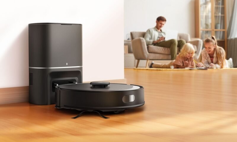 Um robô aspirador de pó preto (TP-Link Tapo RV30 Max Plus) é mostrado em primeiro plano, posicionado na frente de uma estação de acoplamento preta vertical. A estação tem uma pequena luz branca na frente. O robô aspirador tem um disco cinza no centro e um pequeno disco cinza na lateral. Está sobre um piso de madeira bege claro. No fundo, uma família de três pessoas (um homem sentado em um sofá bege com almofadas cinzas e uma menina com uma garota um pouco mais velha estão deitadas no chão. Todos estão usando roupas casuais. O homem parece trabalhar em um notebook enquanto as meninas parecem se divertir desenhando ou escrevendo. O cômodo é bem iluminado com luz natural e apresenta uma parede branca com batente de porta bege.
