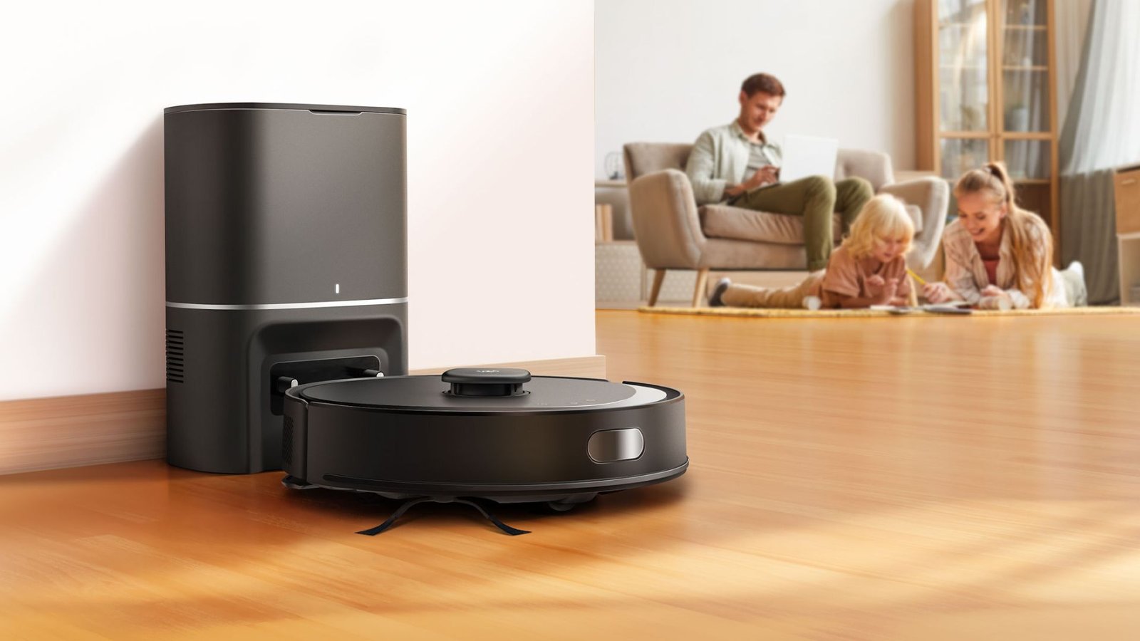 Um robô aspirador de pó preto (TP-Link Tapo RV30 Max Plus) é mostrado em primeiro plano, posicionado na frente de uma estação de acoplamento preta vertical. A estação tem uma pequena luz branca na frente. O robô aspirador tem um disco cinza no centro e um pequeno disco cinza na lateral. Está sobre um piso de madeira bege claro. No fundo, uma família de três pessoas (um homem sentado em um sofá bege com almofadas cinzas e uma menina com uma garota um pouco mais velha estão deitadas no chão. Todos estão usando roupas casuais. O homem parece trabalhar em um notebook enquanto as meninas parecem se divertir desenhando ou escrevendo. O cômodo é bem iluminado com luz natural e apresenta uma parede branca com batente de porta bege.