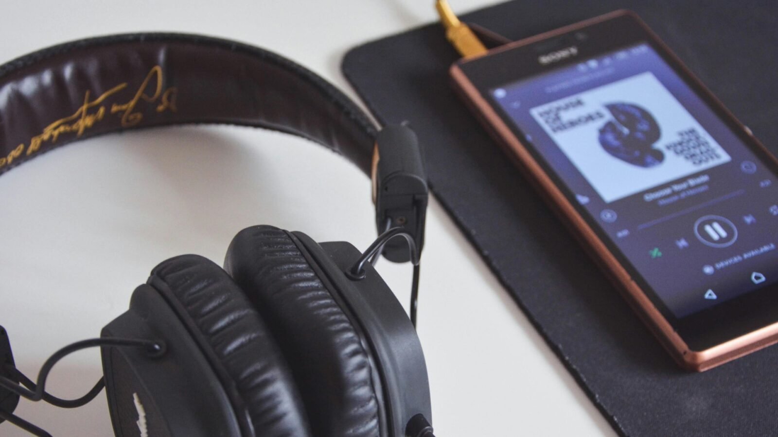 A imagem mostra um par de fones de ouvido Marshall Major II pretos em cima de uma mesa branca, com um smartphone Sony próximo a eles. O smartphone está tocando música em um app de streaming, mostrando a capa do álbum "Heroes" do David Bowie na tela. O fone de ouvido está parcialmente dentro do enquadramento, com o cabo conectado ao smartphone, sugerindo que a música está sendo reproduzida através do fone de ouvido. A mesa tem uma superfície lisa e limpa, e o fundo é levemente desfocado, destacando os fones de ouvido e o smartphone como os focos principais da imagem. A composição da imagem sugere um ambiente relaxante e íntimo, talvez um espaço de trabalho ou um local de descanso. Foto de Mihis Alex