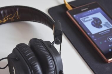 A imagem mostra um par de fones de ouvido Marshall Major II pretos em cima de uma mesa branca, com um smartphone Sony próximo a eles. O smartphone está tocando música em um app de streaming, mostrando a capa do álbum "Heroes" do David Bowie na tela. O fone de ouvido está parcialmente dentro do enquadramento, com o cabo conectado ao smartphone, sugerindo que a música está sendo reproduzida através do fone de ouvido. A mesa tem uma superfície lisa e limpa, e o fundo é levemente desfocado, destacando os fones de ouvido e o smartphone como os focos principais da imagem. A composição da imagem sugere um ambiente relaxante e íntimo, talvez um espaço de trabalho ou um local de descanso. Foto de Mihis Alex