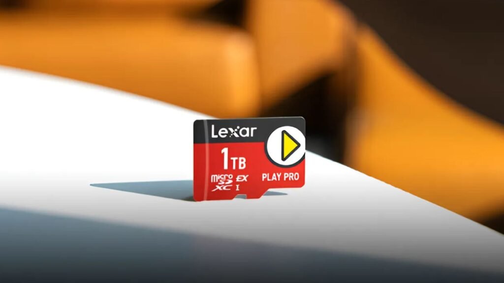 A imagem mostra um cartão de memória microSD vermelho com a marca Lexar, com capacidade de 1TB. O cartão está em cima de uma superfície branca com uma sombra projetada abaixo dele. O fundo está embaçado com uma combinação de cores laranja e branca.