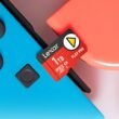 Um cartão de memória microSD vermelho Lexar de 1TB está posicionado acima de dois Joy-Cons azul e vermelho. O texto "Lexar", "1TB", "microSD EX PLAY PRO XC I" e um triângulo amarelo são visíveis no cartão.