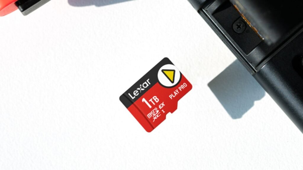 Um cartão de memória microSD vermelho Lexar de 1TB está em cima de uma superfície branca. O texto "Lexar", "1TB", "microSD EX PLAY PRO XC I" e um triângulo amarelo são visíveis no cartão. A parte superior de um Joy-Con vermelho do Nintendo Switch está visível no canto superior esquerdo e próximo ao cartão microSD, é possível visualizar a entrada de cartões de memória do Nintendo Switch aberta para inserção da memória.