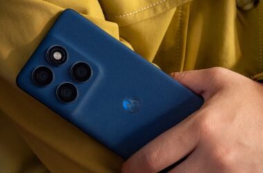 Um homem segura o smartphone azul escuro Motorola Edge 60 5G. A parte traseira do celular está em foco, mostrando o módulo de câmera retangular com três lentes e um sensor/flash. O logo da Motorola aparece na parte inferior. Ao fundo, desfocado, há uma roupa amarelo mostarda.