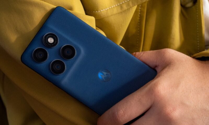 Um homem segura o smartphone azul escuro Motorola Edge 60 5G. A parte traseira do celular está em foco, mostrando o módulo de câmera retangular com três lentes e um sensor/flash. O logo da Motorola aparece na parte inferior. Ao fundo, desfocado, há uma roupa amarelo mostarda.