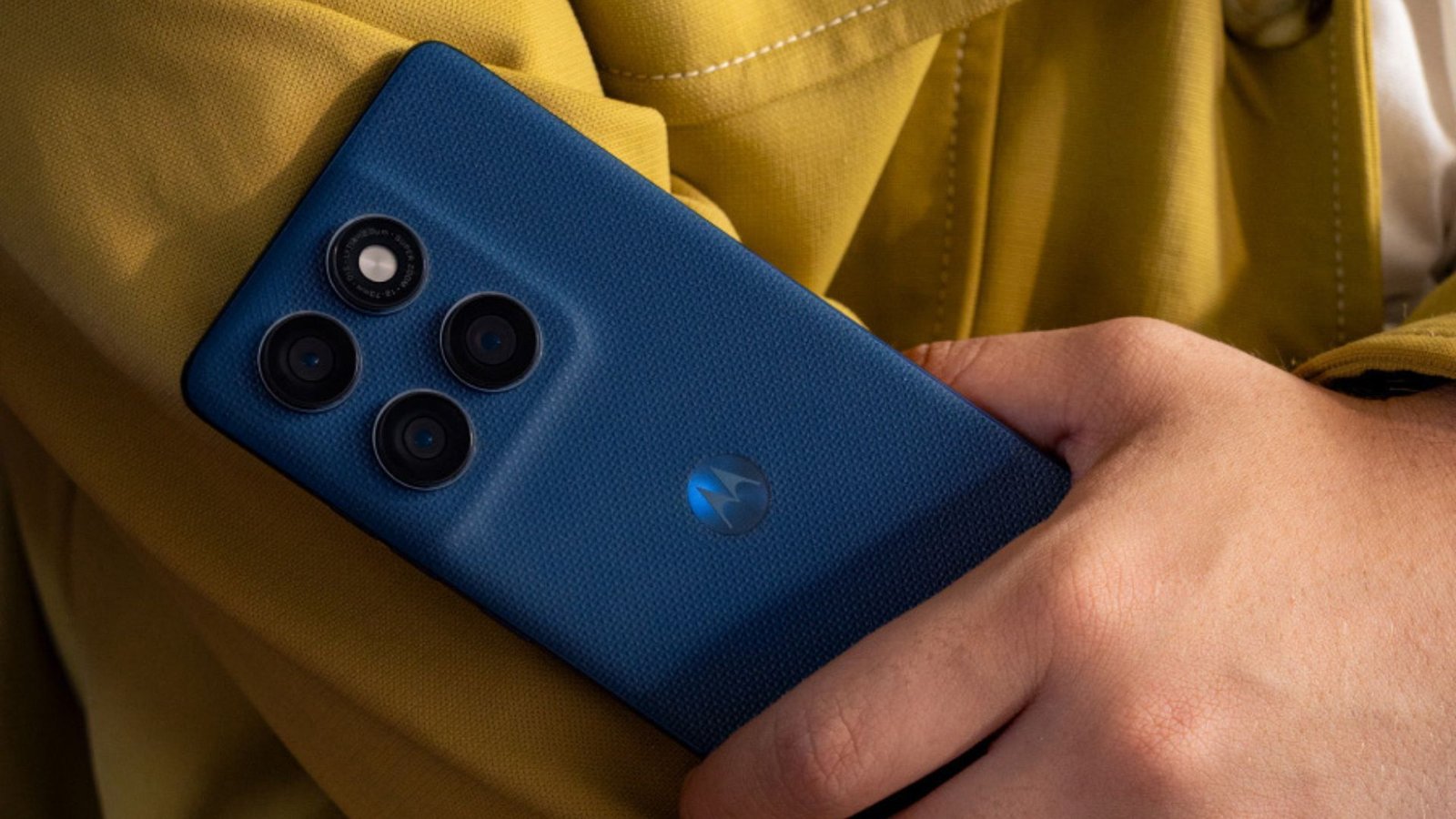 Um homem segura o smartphone azul escuro Motorola Edge 60 5G. A parte traseira do celular está em foco, mostrando o módulo de câmera retangular com três lentes e um sensor/flash. O logo da Motorola aparece na parte inferior. Ao fundo, desfocado, há uma roupa amarelo mostarda.