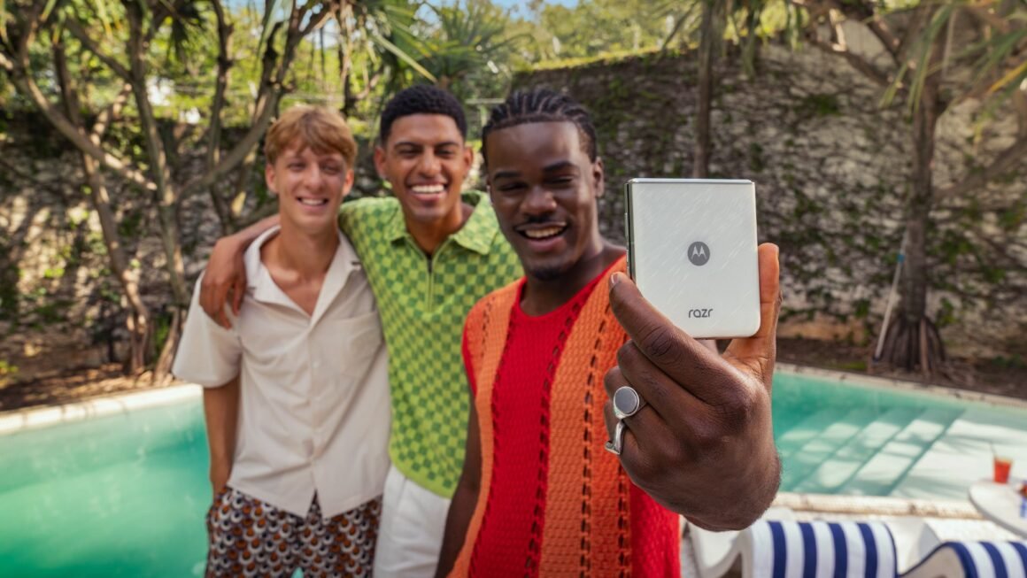 Três jovens adultos sorridentes posam para uma selfie tirada com um smartphone Motorola Razr 60 prateado. O indivíduo mais próximo da câmera, que parece ser um homem, tem pele em um tom mais escuro, usa tranças e veste uma camiseta vermelha e um casaco de malha laranja. Ele segura o celular com a mão direita, onde se destaca um anel grande e claro. À esquerda, outro indivíduo, que aparenta ser um homem com pele clara e cabelos castanhos curtos, veste uma camiseta branca e shorts estampados. No centro, um terceiro indivíduo, também parecendo ser um homem com pele em um tom médio e cabelos curtos e escuros, veste uma camisa verde e branca quadriculada. Os três estão abraçados, demonstrando proximidade e alegria. O fundo da imagem sugere um ambiente de lazer ao ar livre, possivelmente perto de uma piscina com água azul-turquesa e espreguiçadeiras com listras azuis e brancas. Vegetação tropical e uma parede de pedra clara são visíveis ao fundo, indicando um dia ensolarado.
