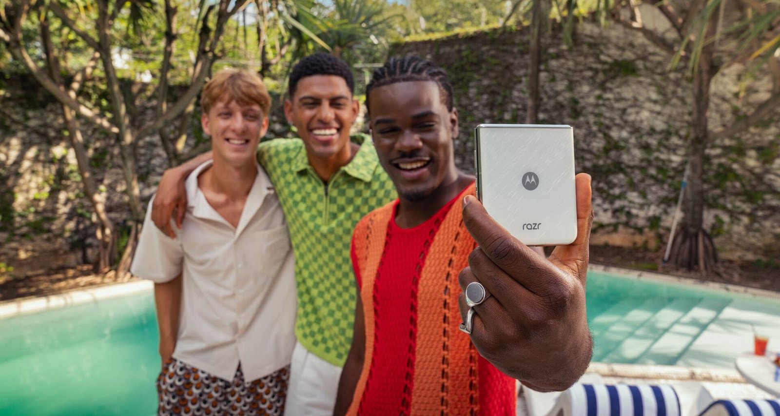 Três jovens adultos sorridentes posam para uma selfie tirada com um smartphone Motorola Razr 60 prateado. O indivíduo mais próximo da câmera, que parece ser um homem, tem pele em um tom mais escuro, usa tranças e veste uma camiseta vermelha e um casaco de malha laranja. Ele segura o celular com a mão direita, onde se destaca um anel grande e claro. À esquerda, outro indivíduo, que aparenta ser um homem com pele clara e cabelos castanhos curtos, veste uma camiseta branca e shorts estampados. No centro, um terceiro indivíduo, também parecendo ser um homem com pele em um tom médio e cabelos curtos e escuros, veste uma camisa verde e branca quadriculada. Os três estão abraçados, demonstrando proximidade e alegria. O fundo da imagem sugere um ambiente de lazer ao ar livre, possivelmente perto de uma piscina com água azul-turquesa e espreguiçadeiras com listras azuis e brancas. Vegetação tropical e uma parede de pedra clara são visíveis ao fundo, indicando um dia ensolarado.