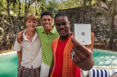 Três jovens adultos sorridentes posam para uma selfie tirada com um smartphone Motorola Razr 60 prateado. O indivíduo mais próximo da câmera, que parece ser um homem, tem pele em um tom mais escuro, usa tranças e veste uma camiseta vermelha e um casaco de malha laranja. Ele segura o celular com a mão direita, onde se destaca um anel grande e claro. À esquerda, outro indivíduo, que aparenta ser um homem com pele clara e cabelos castanhos curtos, veste uma camiseta branca e shorts estampados. No centro, um terceiro indivíduo, também parecendo ser um homem com pele em um tom médio e cabelos curtos e escuros, veste uma camisa verde e branca quadriculada. Os três estão abraçados, demonstrando proximidade e alegria. O fundo da imagem sugere um ambiente de lazer ao ar livre, possivelmente perto de uma piscina com água azul-turquesa e espreguiçadeiras com listras azuis e brancas. Vegetação tropical e uma parede de pedra clara são visíveis ao fundo, indicando um dia ensolarado.