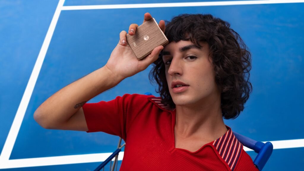 A imagem mostra um indivíduo com cabelos cacheados escuros e pele clara, segurando um smartphone Motorola Razr 60 Ultra na cor bronze/dourado com a mão direita, posicionando-o perto do rosto. A pessoa veste uma camiseta vermelha com detalhes em branco e azul nas mangas e parece estar sentada num carrinho de supermercado azul. O fundo é uma parede azul vibrante com linhas brancas diagonais, sugerindo que o ambiente é uma quadra de tênis (Lisonda). A mão que segura o telefone tem unhas pintadas com diversas cores e desenhos. Uma pequena tatuagem é visível no antebraço esquerdo da pessoa.