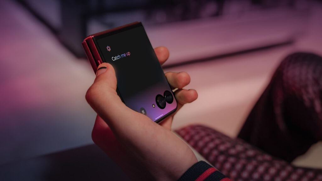 Uma mão segura um smartphone dobrável preto, o Motorola Razr 60 Ultra 5G, fechado. A tela externa exibe a mensagem "Catch me up" com um ícone de balão de diálogo. Ao fundo, desfocado, aparece parte de uma pessoa sentada e um ambiente interno com iluminação suave em tons de rosa e branco.