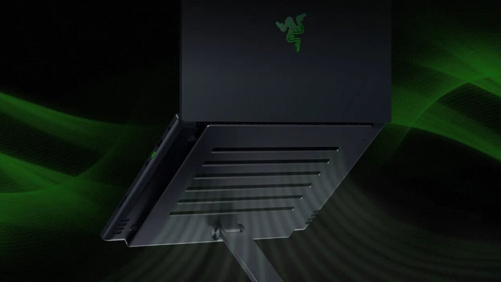 A foto exibe um laptop preto da marca Razer, colocado em um suporte da mesma marca. O suporte apresenta um design ergonômico com ranhuras horizontais, que facilitam a ventilação do laptop e ajudam a mantê-lo resfriado. O fundo da imagem está desfocado, com tons de verde e preto, criando um efeito dramático que destaca o laptop e o suporte. O logo da Razer, em verde neon, é visível na tampa superior do laptop. As ranhuras horizontais no suporte, além de adicionarem um toque estético moderno, também contribuem para a circulação de ar, auxiliando na dissipação de calor gerado pelo laptop.