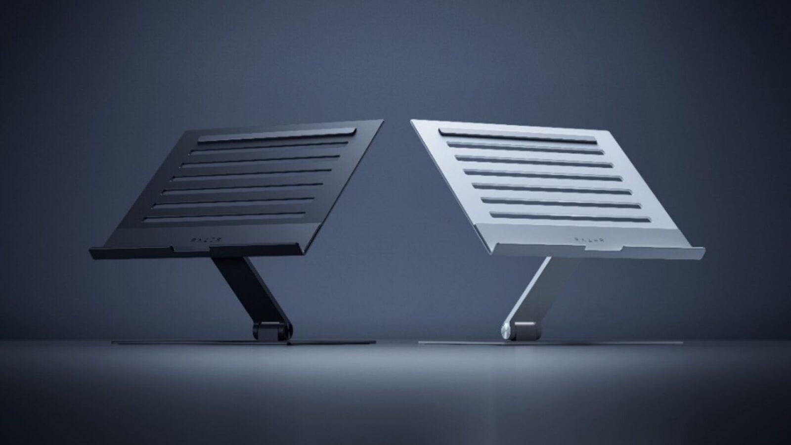 Imagem exibe o Razer Laptop Stand em duas variantes de cor: uma em tom de grafite e outra em tom prateado. Ambas as versões apresentam um design elegante e funcional, com uma base plana e um suporte inclinado para elevar o laptop a uma altura de visualização mais confortável. A superfície do suporte é adornada com ranhuras horizontais para auxiliar na ventilação do laptop. O logo da Razer pode ser visto no canto inferior direito do suporte prateado, indicando a marca do produto.