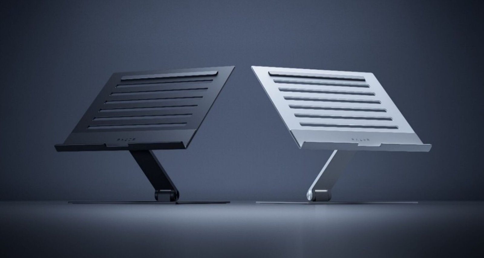 Imagem exibe o Razer Laptop Stand em duas variantes de cor: uma em tom de grafite e outra em tom prateado. Ambas as versões apresentam um design elegante e funcional, com uma base plana e um suporte inclinado para elevar o laptop a uma altura de visualização mais confortável. A superfície do suporte é adornada com ranhuras horizontais para auxiliar na ventilação do laptop. O logo da Razer pode ser visto no canto inferior direito do suporte prateado, indicando a marca do produto.