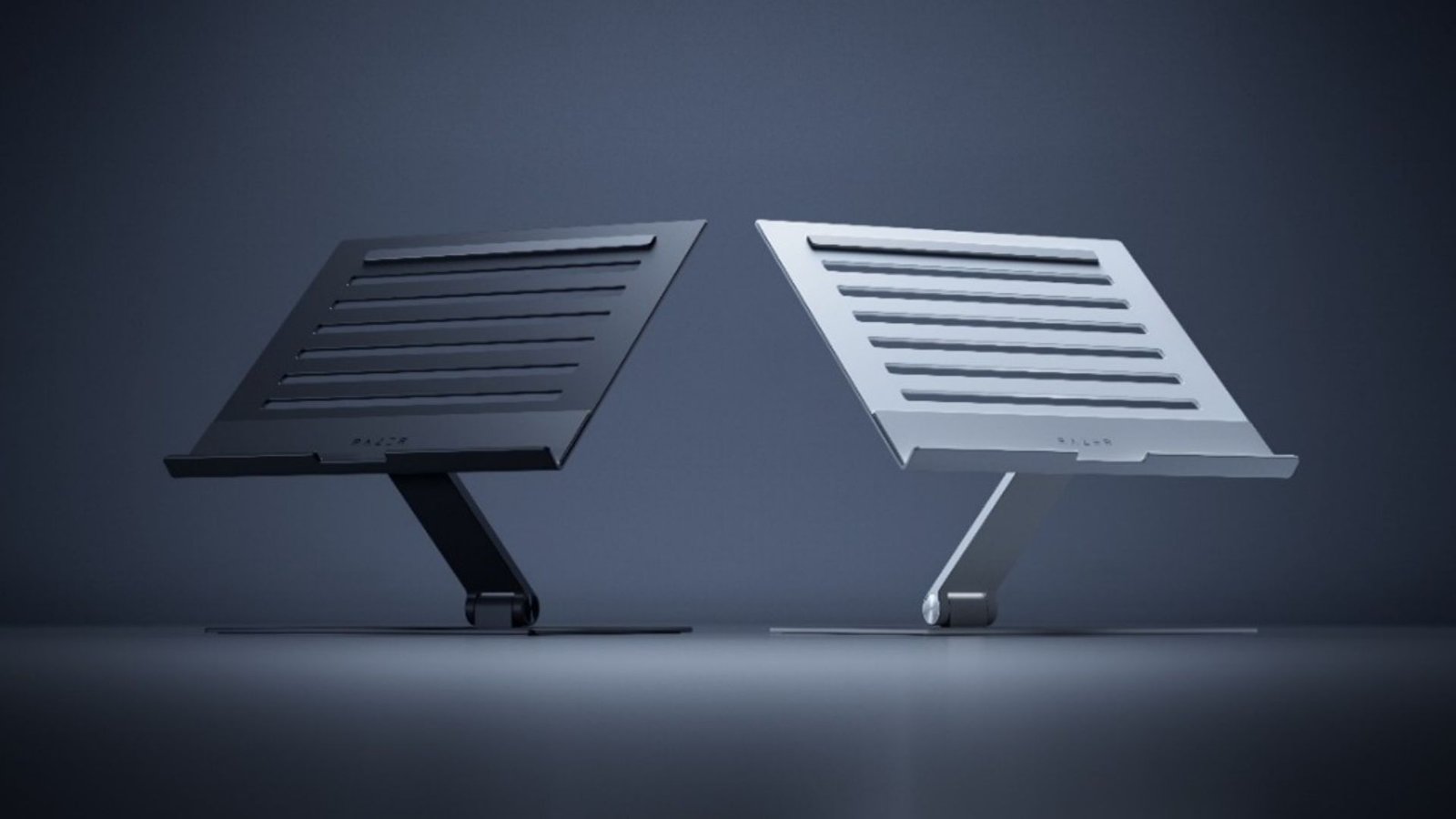 Imagem exibe o Razer Laptop Stand em duas variantes de cor: uma em tom de grafite e outra em tom prateado. Ambas as versões apresentam um design elegante e funcional, com uma base plana e um suporte inclinado para elevar o laptop a uma altura de visualização mais confortável. A superfície do suporte é adornada com ranhuras horizontais para auxiliar na ventilação do laptop. O logo da Razer pode ser visto no canto inferior direito do suporte prateado, indicando a marca do produto.