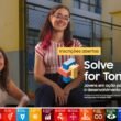 Duas mulheres sorridentes, uma sentada em uma mesa de madeira e outra no banco, em frente a um fundo desfocado com ícones coloridos dos Objetivos de Desenvolvimento Sustentável e o texto em português "Inscrições abertas Solve for Tomorrow Jovens em ação para o desenvolvimento sustentável".