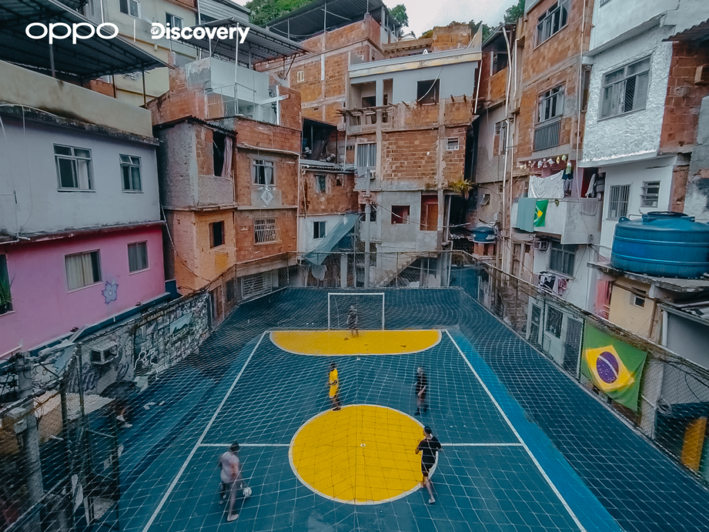Uma fotografia aérea de um campo de futebol em uma favela brasileira. O campo é retangular, com piso azul escuro e dois círculos amarelos no centro. Podem ser visto ao todo cinco pessoas no campo, sendo um goleiro e quatro jogadores de futebol estão espalhados pelo campo. O campo é cercado por uma cerca de rede branca. Ao redor do campo, há uma densa área urbana com casas de tijolos e cores vibrantes, algumas com bandeiras do Brasil penduradas. O logotipo "OPPO" e a palavra "Discovery" são visíveis no canto superior esquerdo da imagem.