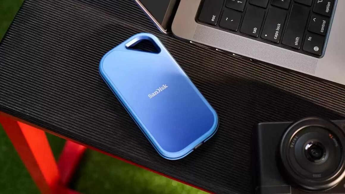 Um SSD portátil SanDisk Creator Pro azul está sobre uma superfície preta texturizada perto de um laptop e uma câmera.