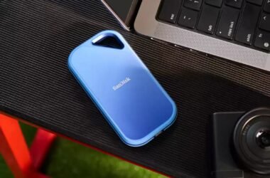 Um SSD portátil SanDisk Creator Pro azul está sobre uma superfície preta texturizada perto de um laptop e uma câmera.