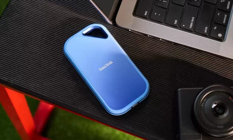 Um SSD portátil SanDisk Creator Pro azul está sobre uma superfície preta texturizada perto de um laptop e uma câmera.