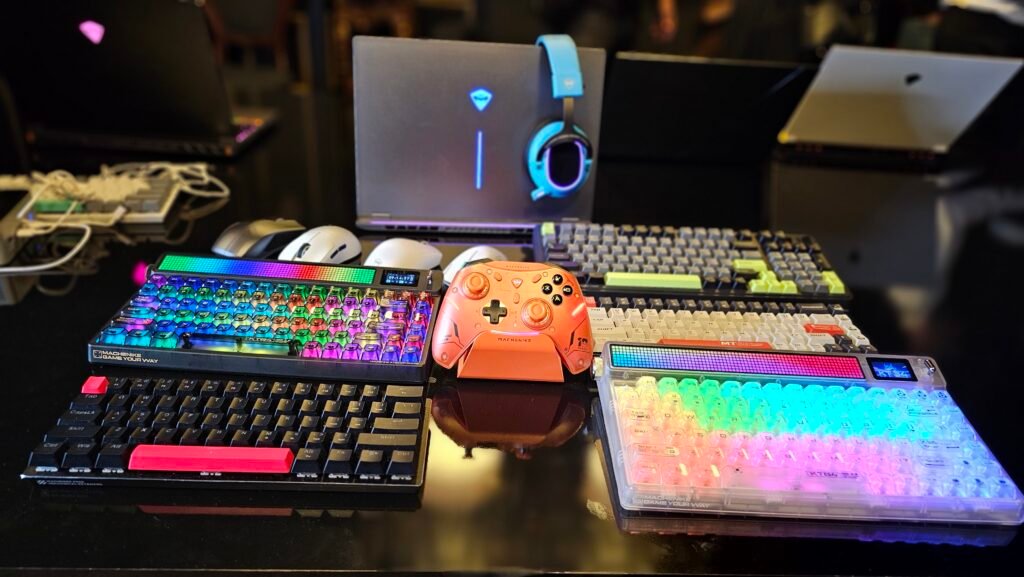 Periféricos de jogos da Machenike em uma mesa escura, com teclados RGB, um controle laranja e um laptop ao fundo, vistos de um ângulo ligeiramente elevado.