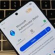 Um smartphone está deitado sobre o teclado preto de um notebook. Na tela do celular, está aberta a página de instalação do aplicativo de senhas "Microsoft Authenticator (Beta)" na Google Play Store. A interface está em português, e o botão azul "Instalar" está em destaque. O teclado ao fundo tem as teclas "option" e "command" parcialmente visíveis.
