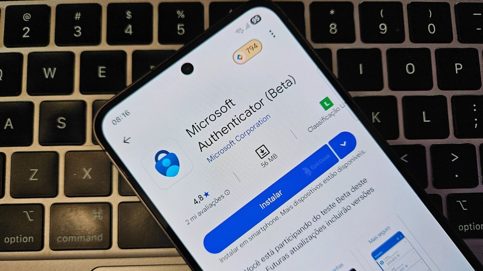 Um smartphone está deitado sobre o teclado preto de um notebook. Na tela do celular, está aberta a página de instalação do aplicativo de senhas "Microsoft Authenticator (Beta)" na Google Play Store. A interface está em português, e o botão azul "Instalar" está em destaque. O teclado ao fundo tem as teclas "option" e "command" parcialmente visíveis.