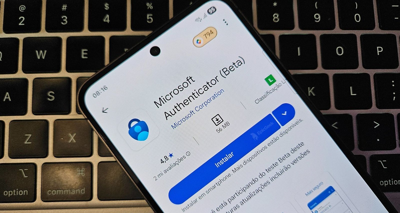 Um smartphone está deitado sobre o teclado preto de um notebook. Na tela do celular, está aberta a página de instalação do aplicativo de senhas "Microsoft Authenticator (Beta)" na Google Play Store. A interface está em português, e o botão azul "Instalar" está em destaque. O teclado ao fundo tem as teclas "option" e "command" parcialmente visíveis.
