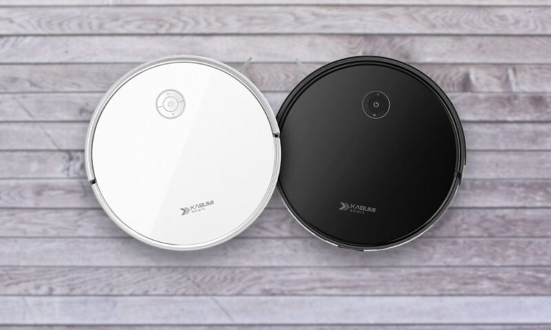 Duas imagens aéreas de aspiradores de pó robôs redondos KaBuM! Smart 550, idênticos em design, mas com cores diferentes. O aspirador à esquerda é branco e o da direita é preto. Ambos possuem um painel de controle simples no topo e a marca "KaBuM! SMART" impressa. Eles estão lado a lado, sobre um piso de madeira clara.
