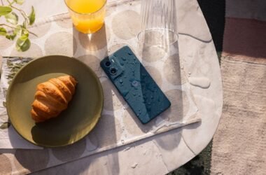 O smartphone Motorola Moto G06 na cor azul marinho, molhado, deitado em uma mesa. Ao lado do aparelho, há um croissant em um prato e um copo de suco.